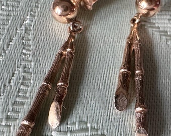 May include: A pair of gold-toned earrings. Each earring features a round stud, a small floral accent, and two bamboo-shaped dangles. The earrings are displayed on a light green textured surface.