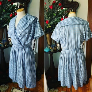 May include: A vintage blue and white gingham dress with a wrap-style bodice and pleated skirt. The dress features a wide collar and short sleeves. The dress is displayed on a mannequin.