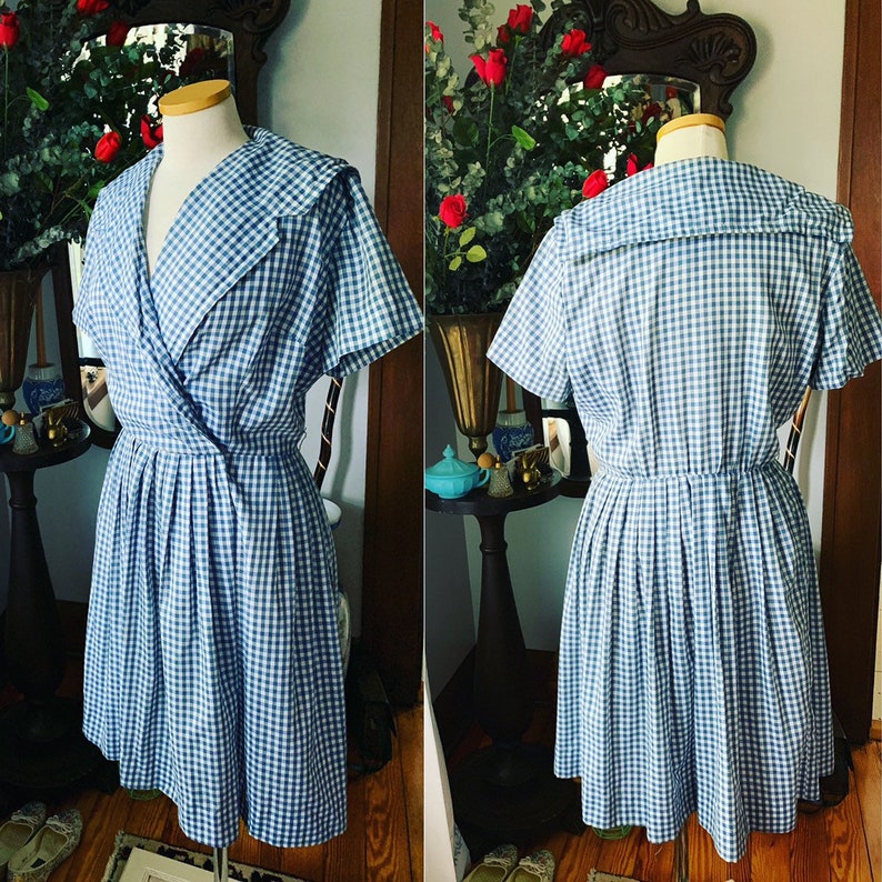 May include: A vintage blue and white gingham dress with a wrap-style bodice and pleated skirt. The dress features a wide collar and short sleeves. The dress is displayed on a mannequin.
