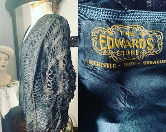 Può includere: Una giacca nera con apertura frontale e dettagli traforati intricati. La giacca è esposta su un manichino. L'etichetta Edwards Stores è visibile, con testo che indica le sedi a Rochester, Troy e Syracuse. Il design della giacca presenta un motivo tessuto.