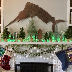 May include: A white fireplace mantel decorated for Christmas with a garland of greenery and white pom poms. The garland has a banner that reads "Merry Christmas". There are small potted evergreen trees and a metal marlin sculpture on the mantel. There are also three Christmas stockings hanging on the mantel.