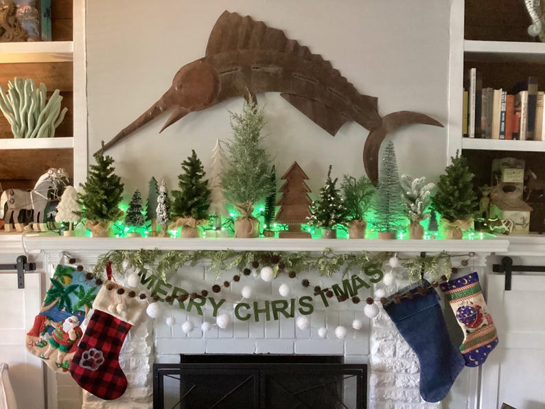 May include: A white fireplace mantel decorated for Christmas with a garland of greenery and white pom poms. The garland has a banner that reads "Merry Christmas". There are small potted evergreen trees and a metal marlin sculpture on the mantel. There are also three Christmas stockings hanging on the mantel.