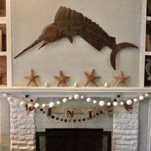 May include: A white brick fireplace with a white garland of pom poms and a burlap banner that reads "Winter". Above the fireplace is a metal marlin sculpture and four starfish.