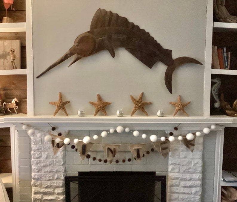 May include: A white brick fireplace with a white garland of pom poms and a burlap banner that reads "Winter". Above the fireplace is a metal marlin sculpture and four starfish.