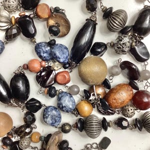 May include: A collection of necklaces with various beads in black, brown, gray, and blue. The beads include wooden, metal, and stone elements, strung on silver-colored chains. Some beads have a ribbed or textured design.