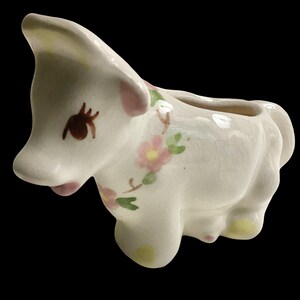 May include: A ceramic donkey-shaped creamer with a white base, adorned with pink and green floral accents. The donkey has brown eyes and pink accents on its ears and snout. The creamer has a small opening at the top for pouring.