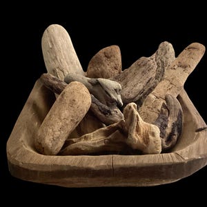 May include: A rectangular wooden dough bowl filled with a collection of weathered driftwood pieces. The bowl features a natural, light brown wood grain, while the assorted driftwood varies in shape, size, and texture, displaying shades of tan, gray, and dark brown.