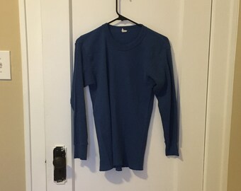 May include: A long-sleeved, blue thermal shirt hangs on a black hanger against a white door. The shirt has a ribbed texture and a crew neck. The cuffs and hem are finished with a simple edge.