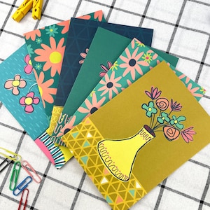 May include: A selection of floral-themed greeting cards. Each card displays a unique design, including flowers, geometric patterns, and a vase of flowers. The cards are arranged in a fan shape, showcasing their vibrant colours and artistic illustrations.