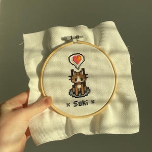 Peut inclure: Un cerceau de broderie blanc avec un motif de point de croix représentant un chat brun avec un cœur rouge au-dessus. Le chat est assis sur une surface à motifs bleus et blancs. Le texte "x Suki x" est brodé sous le chat.