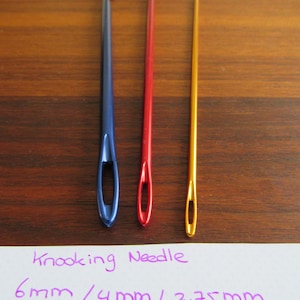 Può includere: Tre uncinetti, uno blu, uno rosso e uno dorato, con un cartoncino bianco che dice "Crooking Needle 6mm / 4mm / 2.75mm".
