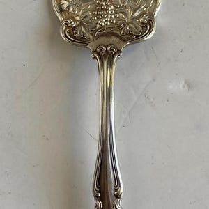 May include: A silver fork with an ornate handle featuring a grapevine design. The fork has four tines and a slightly bent handle.