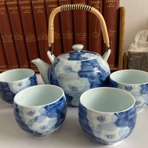 May include: A blue and white floral patterned tea set, including a teapot with a woven handle and four matching teacups. The tea set is made of ceramic and is displayed on a white surface. The background includes a collection of books.