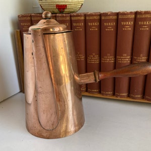 May include: A copper teapot with a wooden handle. The teapot is sitting on a shelf with a row of books behind it. The books are all brown and have gold lettering on the spines. The books are titled "Shakespeare's Works".