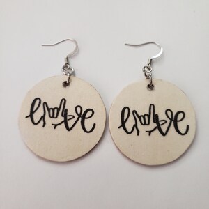 May include: A pair of wooden earrings with the word "LOVE" spelled out in American Sign Language. The earrings are round and have a cream color.