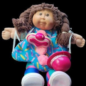 Könnte beinhalten: Eine Vintage-Puppe mit braunen Garnhaaren, einem runden Gesicht und einem blauen Kleid mit farbenfrohem Muster. Die Puppe hat ein Zubehör im Clown-Stil mit einem rosa Hut und einem rosa-weißen Clownkragen. Die Schuhe der Puppe sind weiß mit rosa Akzenten.