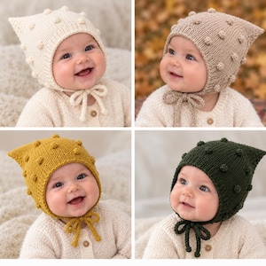 May include: A collection of four knitted baby bonnets featuring a pointed top and small bobble accents. The bonnets are available in cream, beige, mustard yellow, and dark green, each with matching knitted chin ties for secure fastening.