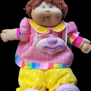 Könnte beinhalten: Eine Vintage-Cabbage-Patch-Kids-Puppe, die als Clown verkleidet ist. Die Puppe hat ein rundes Gesicht, blaue Augen und braunes Garnhaar. Sie trägt ein rosa kariertes Kleid mit gelbem Kragen, gelbe Punktehose und ein Clown-Gesicht-Accessoire.