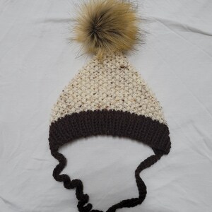 May include: A knitted winter hat with a brown brim and ear flaps. The hat is cream-coloured with brown flecks and a large, fluffy, light brown pom-pom on top. The ear flaps are dark brown.
