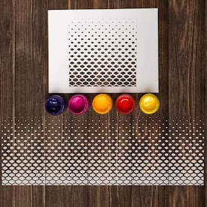 May include: A stencil with a repeating pattern of hexagons, creating a gradient effect. The stencil is white and is on a wooden surface. There are five small paint pots in different colors: purple, pink, yellow, red, and orange.