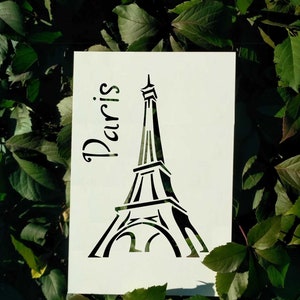 May include: A white stencil featuring the Eiffel Tower and the word "Paris" in a script font. The black silhouette of the tower is set against a background of green leaves.