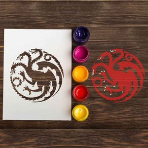 Puede incluir: Una plantilla blanca del sello de la Casa Targaryen, un dragón de tres cabezas, con una versión pintada de rojo del sello a la derecha. La plantilla está sobre una superficie de madera con cinco botes de pintura de varios colores.