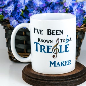 Puede incluir: Taza de cerámica blanca con el texto "I've Been Known to be a Treble Maker" en negro y azul. La taza está sobre un posavasos marrón oscuro. Una cesta de flores azules y blancas está en el fondo.