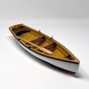 Puede incluir: Un pequeño bote de remos de madera con casco blanco e interior amarillo. El bote tiene dos remos de madera y está sobre una superficie blanca.