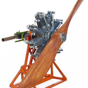 May include: A model airplane engine with a wooden propeller. The engine is silver and has multiple cylinders. The propeller is a light brown color. The engine is mounted on an orange metal stand. A brass tube extends from the engine.