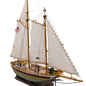 May include: A detailed model sailboat with white sails, wooden masts, and a green and yellow hull. An American flag is visible on the side of the boat. The model is mounted on a dark wooden base.