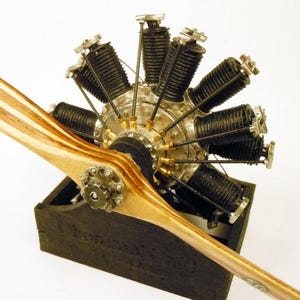 May include: A detailed model of an aircraft engine with a wooden propeller. The engine features multiple cylinders and intricate metal components. The propeller is light brown with a logo. The engine is mounted on a black base.