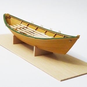 May include: A wooden model boat with a yellow hull and green trim, resting on a light brown base. The boat contains several wooden oars. The model is displayed on a light brown rectangular base.