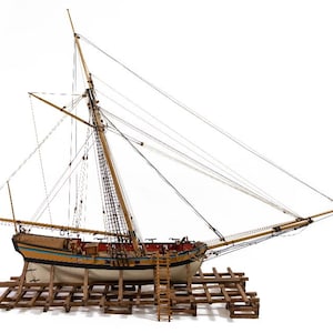 May include: A detailed model sailboat with tan masts, rigging, and a wooden hull. The hull features yellow, blue, and red accents. The model is mounted on a wooden cradle with a small ladder, showcasing nautical craftsmanship.