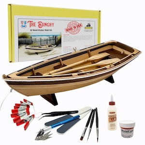Puede incluir: Un kit de modelo de madera de un bote dinghy con remos, exhibido en un soporte. El kit incluye herramientas, pegamento y una caja con el texto "The Dinghy All Wood Display Model Kit" y "Made in USA".