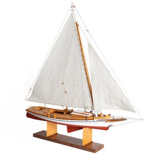 May include: A detailed model sailboat with white sails and a wooden hull. The boat is mounted on a wooden stand, showcasing its intricate design. The hull features red and white accents, and the sails are fully deployed.