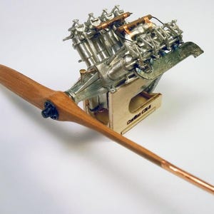 May include: A detailed model of an aircraft engine and propeller. The engine is silver with copper-colored accents, mounted on a wooden stand. The propeller is made of wood with a copper tip. The model is set against a white background.