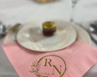 May include: A light pink paper napkin featuring a gold foil printed design. The design includes a circular frame with a leafy branch on the left side, the letters R and N in the center, and the date 03.08.24 printed below the letters.