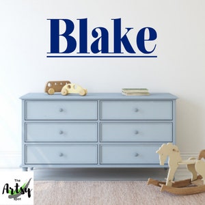 May include: A blue wall decal with the name "Blake" in a bold, stylized font. The decal is above a light blue dresser with six drawers and silver knobs. A wooden toy bus and car are on top of the dresser, and a rocking horse is in front of the dresser.