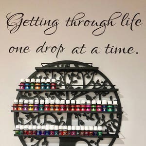 May include: A black metal essential oil display shelf in the shape of a tree, filled with numerous small bottles of essential oils. The wall above the shelf has the text "Getting through life one drop at a time."
