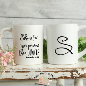 May include: Two white ceramic mugs. One mug features the text "She is far more precious than JEWELS. Proverbs 31:10" in black script. The other mug has a black script letter "S". Both mugs have a curved handle.