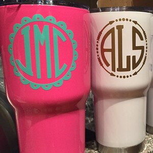 May include: Two stainless steel tumblers with personalized monograms. One is hot pink with a teal monogram in a scalloped circle. The other is white with a gold monogram in a circle with arrows.