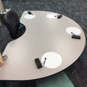 May include: A half-circle table with a light gray surface and four white circles. Each circle has a black marker and a black eraser. The table is in a classroom setting with a blue and green playhouse in the background.