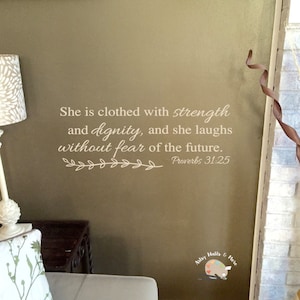May include: A wall decal with a beige background and a white inscription that reads "She is clothed with strength and dignity, and she laughs without fear of the future. Proverbs 31:25".