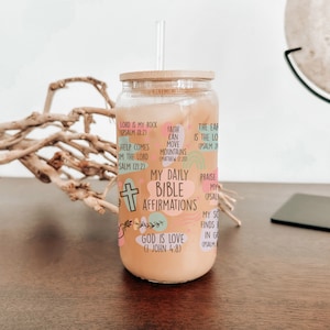 May include: A clear glass tumbler with a bamboo lid and a straw. The tumbler has a colorful design with bible verses and affirmations. The text includes "My Daily Bible Affirmations", "God is Love (I John 4:8)", "Faith can move mountains (Matthew 17:20)", and "Lord is my rock (Psalm 18:2)".