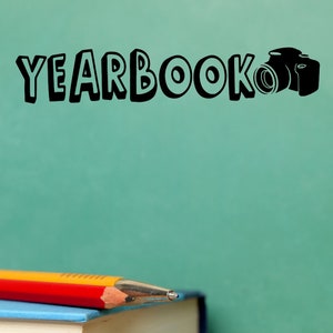 May include: A yellow and red pencil rests on top of a blue and beige book. The word "YEARBOOK" and a camera icon are written in black on a green chalkboard.