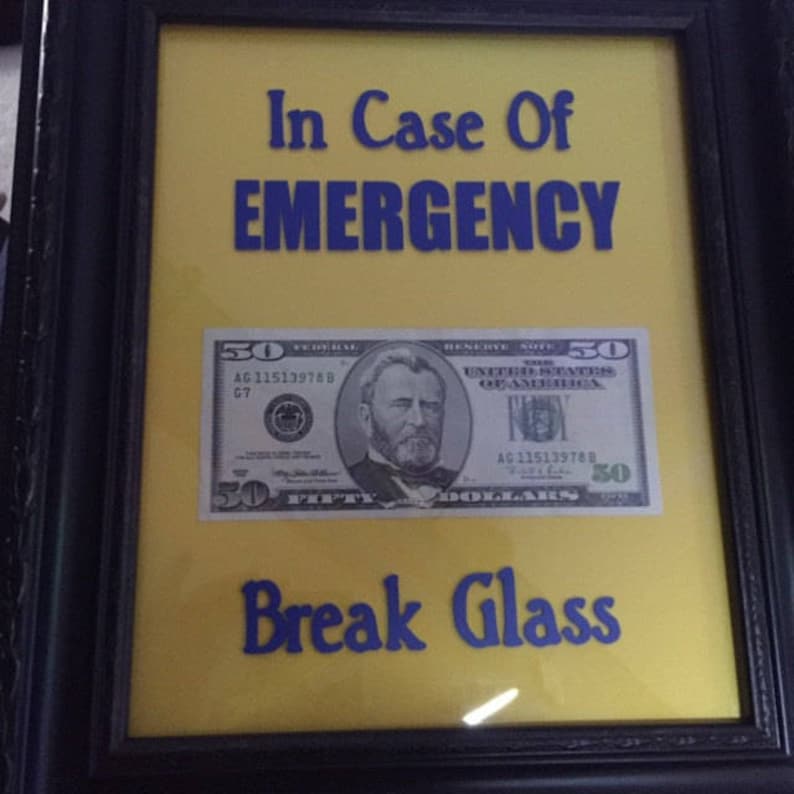 May include: A framed sign with a yellow background and blue text that reads "In Case Of EMERGENCY Break Glass". A $50 bill is behind the glass.