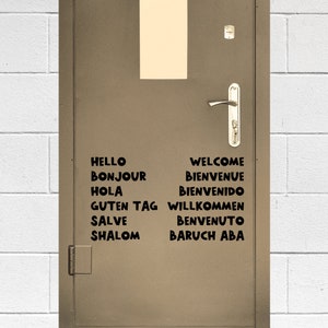 May include: A brown metal door with a silver door handle and a white rectangular sign above the handle. The door has black lettering with the words "Hello", "Bonjour", "Hola", "Guten Tag", "Salve", "Shalom", "Welcome", "Bienvenue", "Bienvenido", "Willkommen", "Benvenuto", and "Baruch Aba" written on it.