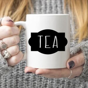 K&ouml;nnte beinhalten: Wei&szlig;e Keramiktasse mit einem schwarzen Etikett, auf dem in einfacher Schrift "TEA" steht.