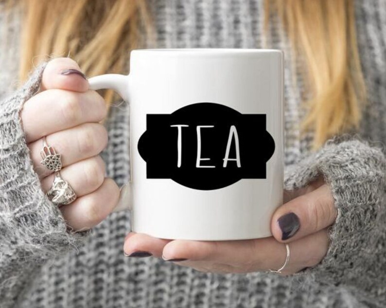 K&ouml;nnte beinhalten: Wei&szlig;e Keramiktasse mit einem schwarzen Etikett, auf dem in einfacher Schrift "TEA" steht.