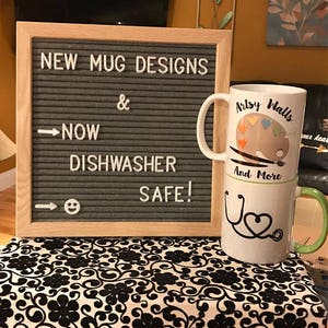 May include: A sign that reads "NEW MUG DESIGNS & NOW DISHWASHER SAFE!" with two mugs stacked on top of each other. The top mug has a painting palette design with the text "Artsy Walls" and the bottom mug has a stethoscope design with the text "And More".